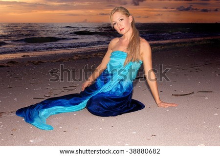 a pretty blonde woman is sitting on the beach at sunset looking at you the viewer