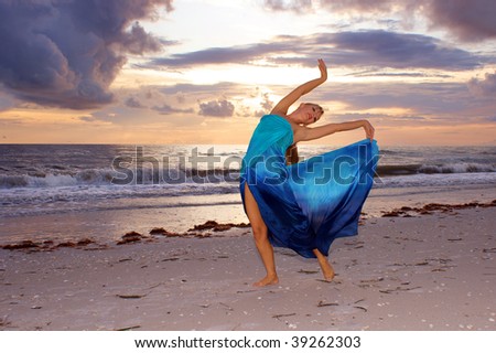 A beautiful blonde ballerina is arching back with her arms up on the beach as the sun sets looking at you the viewer and smiling.