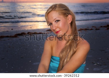 A beautiful blonde young lady is looking at viewer while sitting at the beach while the sunsets in this head and shoulders portrait.