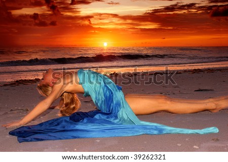 A long haired blonde woman is stretched out on the beach at sunset in profile with her legs straight out and her back arched as she looks up at the sky.