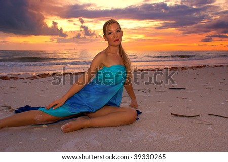 A vibrant tropical sunset over the ocean with an attractive blonde haired woman stretched out on the beach looking directly at viewer.