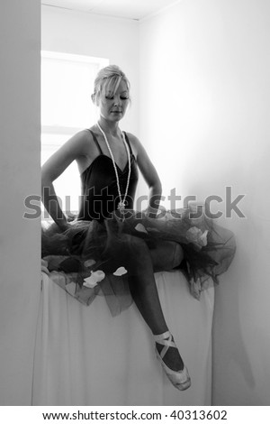 A ballerina is in full costume, sitting o n a window seat with her hands  on her hips and looking down at her feet in this black and white image.