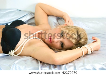 attractive blonde woman is laying on satin sheets wearing a bra with her hands above her head looking seductively at viewer.