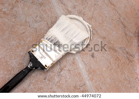 A trim paint brush covered with paint is resting on paint covered cap on floor.