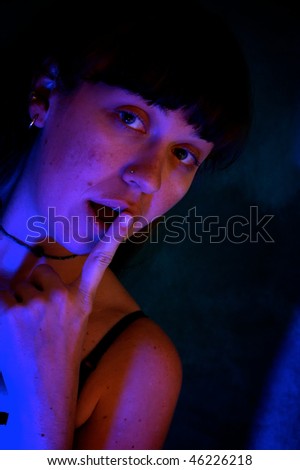 a surprised looking beautiful young woman with wide eyes and finger covering her open mouth in a dark room, lit with red and blue lights