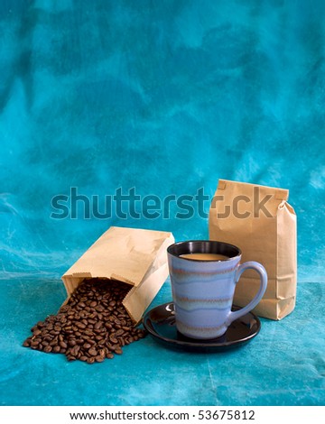 a fresh cup of coffee  in a blue cup with saucer, with coffee beans pouring out of bag and a blank bag standing, add your logo. Also copy space above on mottled blue background