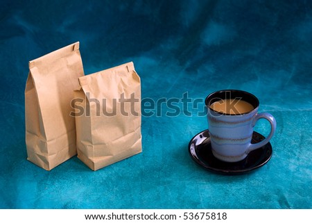 a fresh cup of coffee  in a blue cup with saucer, with two blank bags standing, add your logo. Also copy space above on mottled blue background