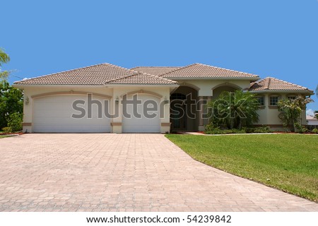 front view a generic florida home sits on a small grass covered hill with garage, palm trees and a clear blue sky, looking up the driveway.