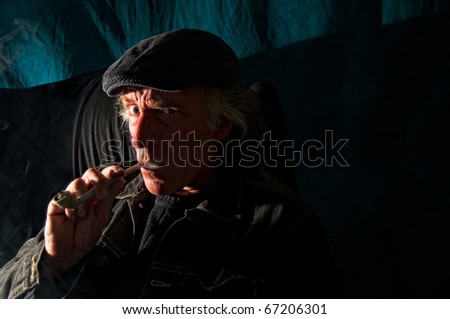 A dangerous looking man with an evil expression on his face is sitting in a dark room looking at viewer smoking pipe.