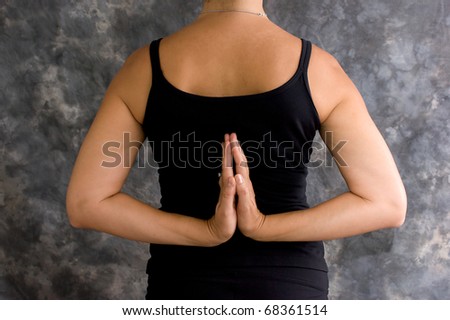 A brunette caucasian woman is standing in black yoga clothing with her hands behind her back in reverse prayer