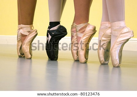Four ballerinas are standing on their toes, on pointe, wearing ballet shoes of various types during dance class.