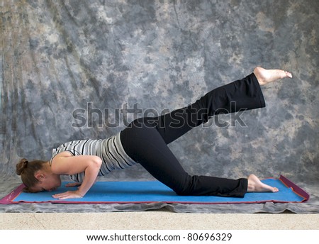 Young woman on yoga mat in  Yoga posture sunbird pose with right leg lifted, against a grey background in profile, facing left lit by diffused sunlight.