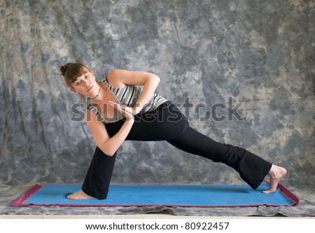 Young woman on yoga mat Yoga posture Parivrtta Parsvakonasana or Revolved Extended Side Angle pose with hands in prayer position against a grey background, facing left lit by diffused sunlight.