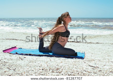 athletic woman is doing yoga exercise on beach in Kapotasana or Pigeon Pose both arms back holding foot on an empty beach at the gulf of mexico in bonita springs florida with long hair blowing in wind