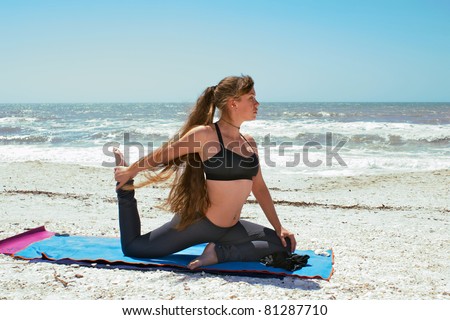 an athletic brown haired woman is doing yoga exercise  on beach in Kapotasana or King Pigeon Pose on an empty beach at the gulf of mexico in bonita springs florida with long hair blowing in wind