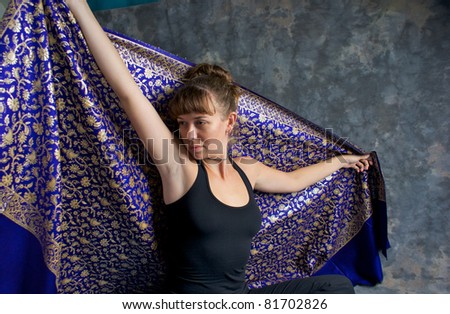 A beautiful young woman dressed in black with head turned to side is holding a shiny large blue and gold cloth behind her with outstretched arms.