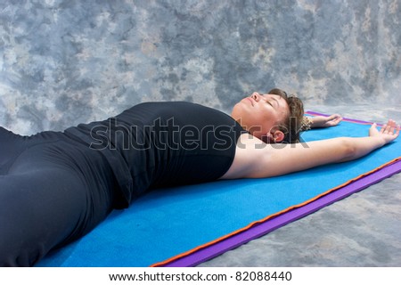 an athletic brown haired woman is stretched out on yoga mat with hands above head in studio with a mottled grey background.