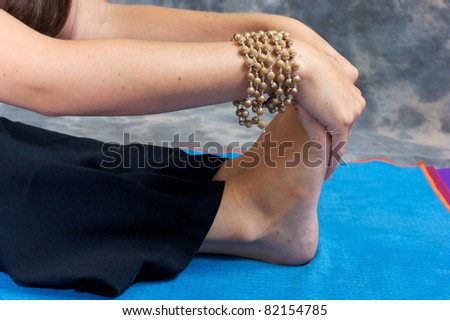 Close up of a woman's hands and feet as she bends over in a yogic forward fold or Paschimottasana on yoga mat in studio wearing mala beads.