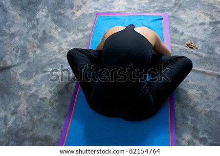 Looking down on an athletic brown haired woman from behind doing yoga exercise Bound Angle Forward Bend pose on yoga mat in studio with mottled background.