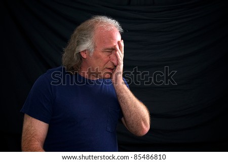 A white man is holding his head as if in despair, he looks upset or hurt against a black background and wearing a t shirt.