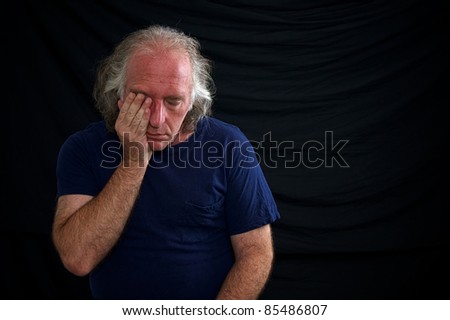 A white man is rubbing his eye and looks sad against a black background and wearing a t shirt.