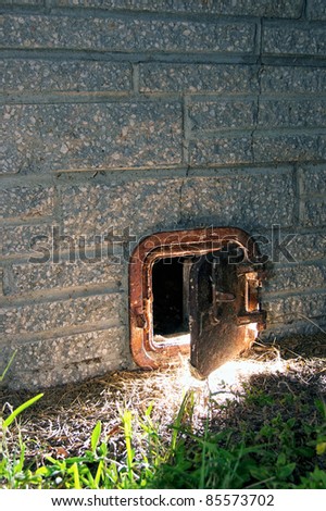 A partially open old rusted iron door sits close to the ground on the outside wall of a stone brick chimney, lit from the inside in the nighttime.