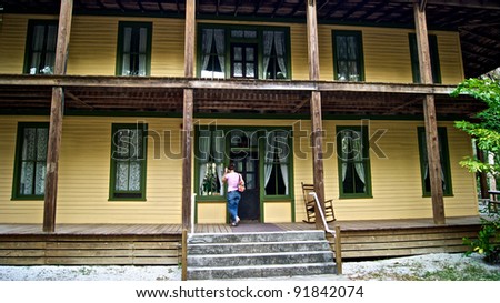 A young woman stands on the porch of an old wooden house built in early 1900\'s in historic area of estero florida. There is an old wooden rocking chair on porch. Koreshan state Park.