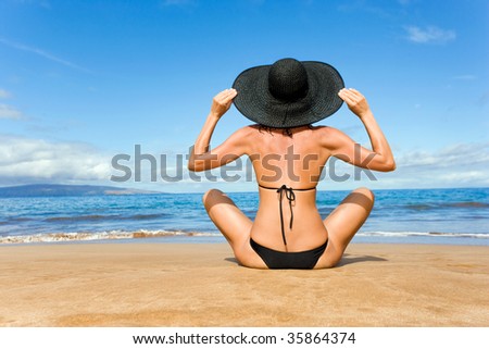 elegant classy woman in black bikini sitting on tropical beach with wide brim hat on vacation in maui, hawaii