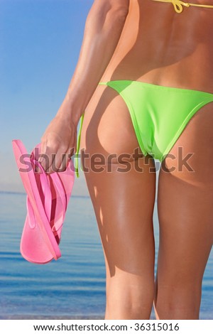 woman holding pink flip flops on tropical beach in green bikini