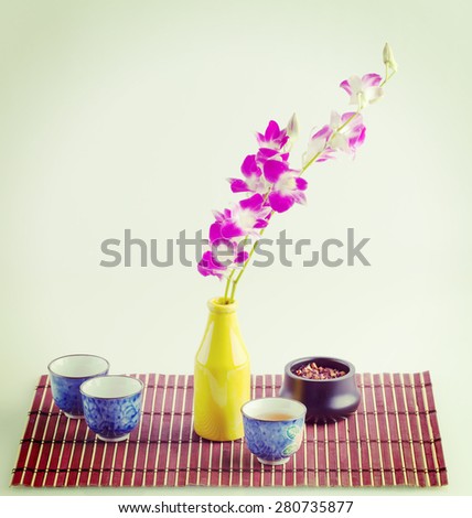 Purple orchid in a yellow bottle and black tea in small Japanese cups on a bamboo mat still life composition.