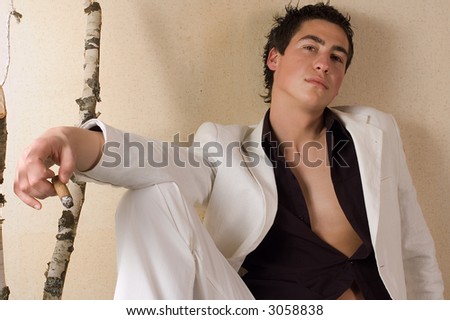 Young male fashion model dressed in white clothes sitting at the desk and holding cigarette