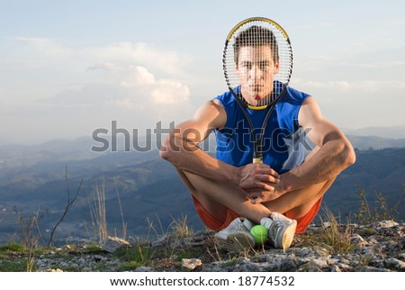Young man holding racket tennis in front of his face, sitting on rock