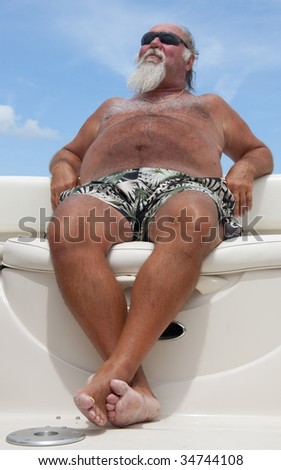 Older man relaxes on a summer day sitting on a boat with a blue sky in the background.