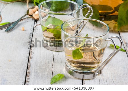 Basil herbal tea in a transparent cup and teapot, spoon with cane sugar, a bunch of fresh basil. On a white wooden table. Copy space