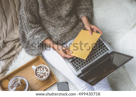 Girl in beige knit sweater and white pants sitting at home in cozy atmosphere and is holding golden envelope.In lap of girl is laptop. Girl using gadget. In the background white knitted pillow.