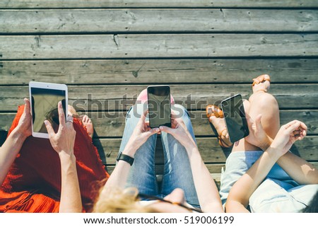 Summer sunny day. Top view, computer tablet and smart phones in the hands of the three girls sitting outside on a wooden platform. Young women using digital gadgets