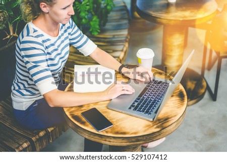 Young woman in striped t-shirt sitting at a round wooden table in cafe and using a laptop. On the table is a notebook, smatrfon and cup of coffee. Online training, interview. Girl uses the gadget.