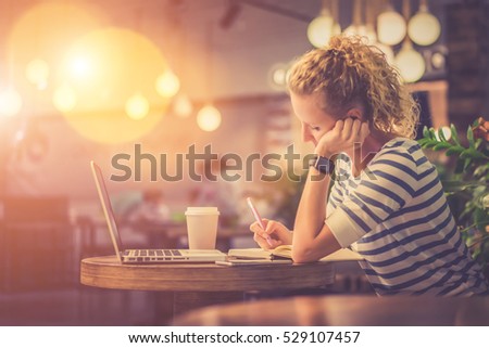 Side view. Young woman in striped blouse sitting at table in cafe and makes notes in notebook. In front of her is laptop next to cup of coffee. Online learning. Student doing homework. Film effect.