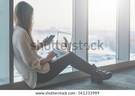 Side view of young businesswoman in white shirt sitting on floor near window and uses laptop and smartphone. In background is blurred cityscape. Student learning online.Girl shopping online, blogging.
