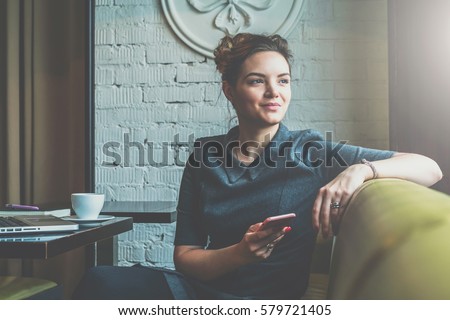 Young smiling business woman sitting in cafe at table, leaning his hand back in chair,looking out window and holding smartphone.On table is laptop, notebook and cup of coffee. Student learning online.