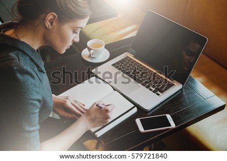 Young business woman in gray dress sitting at table in cafe and writing in notebook. On table is laptop, smartphone and cup of coffee. Freelancer working in coffee shop. Student learning online.