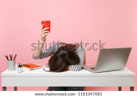 Exhausted woman laid her head down on the table holding cup of coffee or tea sit, work at white desk with pc laptop isolated on pastel pink background. Achievement business career concept. Copy space