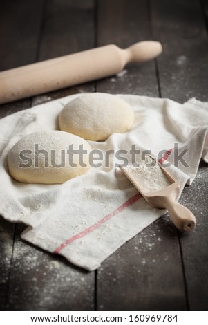 Two floured balls of uncooked homemade pizza dough on a kitchen towel on a rustic dark wooden table with flour and rolling pin.