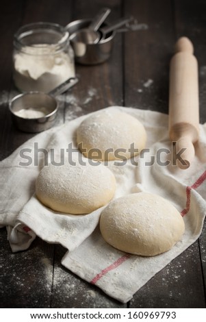 Three floured balls of uncooked homemade pizza dough proofing on a kitchen towel. Taken on a rustic dark wooden table with flour and rolling pin.