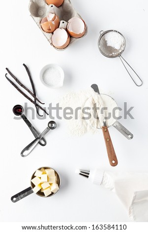 Assorted baking ingredients and tools: eggshells, vanilla pods, flour in measuring cup, spatula, measuring spoon with vanilla extract, butter, sea salt, piping bag and vintage sifter with icing sugar.