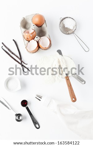 Assorted baking ingredients and tools: eggshells, vanilla pods, flour with spatula in measuring cup, measuring spoons with vanilla extract, sea salt, piping bag and vintage sifter with icing sugar.