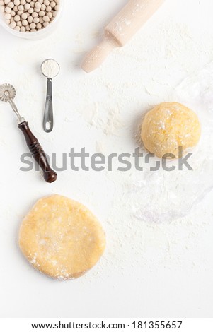 Two unrolled and unbaked shortcrust pastry dough with assorted baking tools: measuring spoon, rolling pin, ceramic baking beans and pastry wheel. Taken on a floured white surface, directly from above.