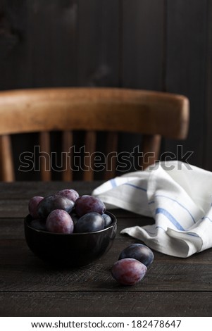 Ripe damson plums served in a black bowl on a black rustic wooden table with a french kitchen towel. A black wood plank wall and an antique chair in the background.