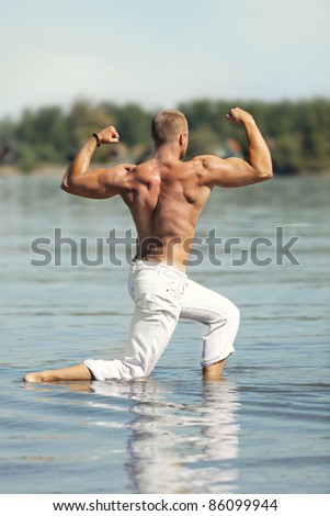 Young male fitness model standing in a shallow river, in a back double biceps bodybuilding pose. Wearing white pants. Shallow depth of field.