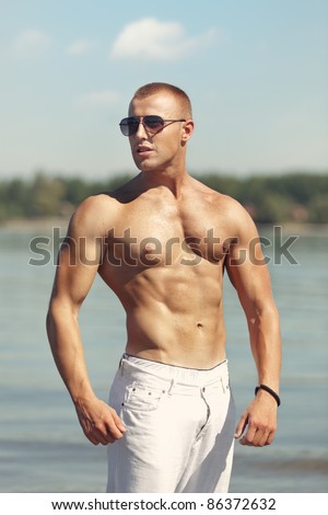 Young male fitness model standing in a river, in a bodybuilding pose. Wearing white pants and sunglasses. Shallow depth of field.
